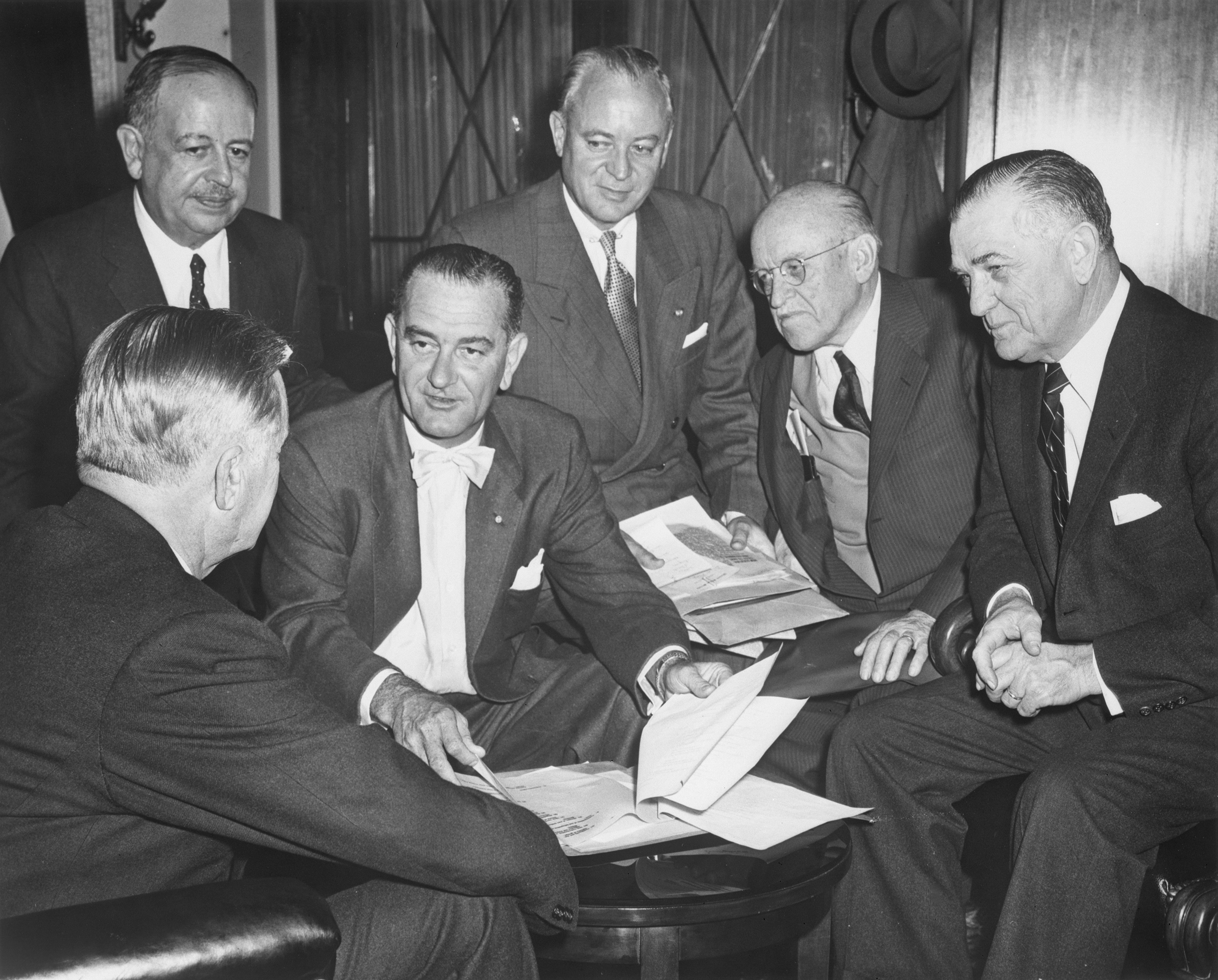Six men in suits in conversation. LBJ (center) looks through some papers.
